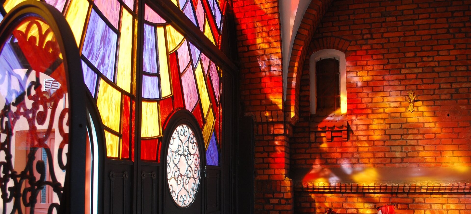 Widok bramy z kolorowymi szybami, przez które pada światło na sąsiednią, ceglaną ścianę.